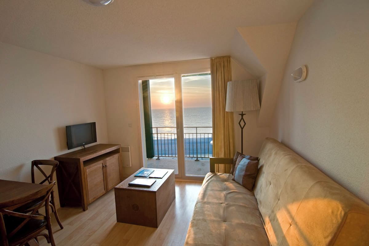 Ein Wohnzimmer mit einer Couch, einem Fernseher auf einer Holzkommode, einem Esstisch mit Stühlen und Blick auf das Meer durch eine Glastür.