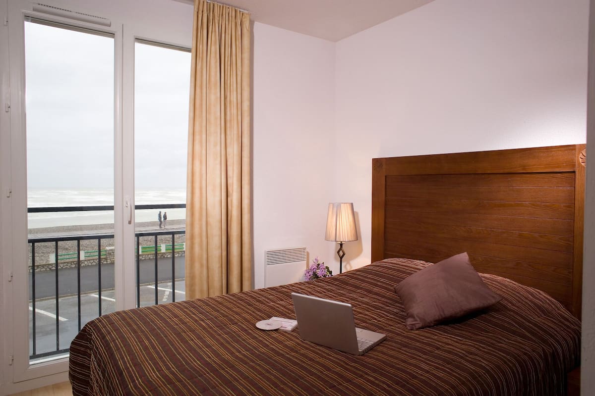 Ein Hotelzimmer mit einem Bett, einem Laptop, einer Lampe und Blick auf den Strand durch das Fenster.