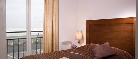 A hotel room with a bed, a laptop, a lamp, and a view of the beach through the window.