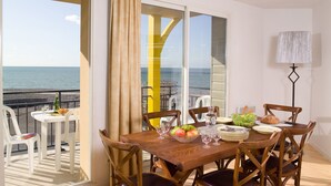 A dining area with a table set for four, a bowl of fruit, and a view of the sea through large windows. - Madame Vacances Residence Les Terrasses de la Plage (Cayeux-sur-Mer)