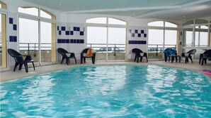An indoor swimming pool with clear blue water, surrounded by black lounge chairs and large windows offering views of parked cars outside. - Madame Vacances Residence Les Terrasses de la Plage (Cayeux-sur-Mer)
