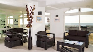 A modern lounge area with wicker furniture, a tall vase with dried leaves, and large windows offering a view of the sea. - Madame Vacances Residence Les Terrasses de la Plage (Cayeux-sur-Mer)