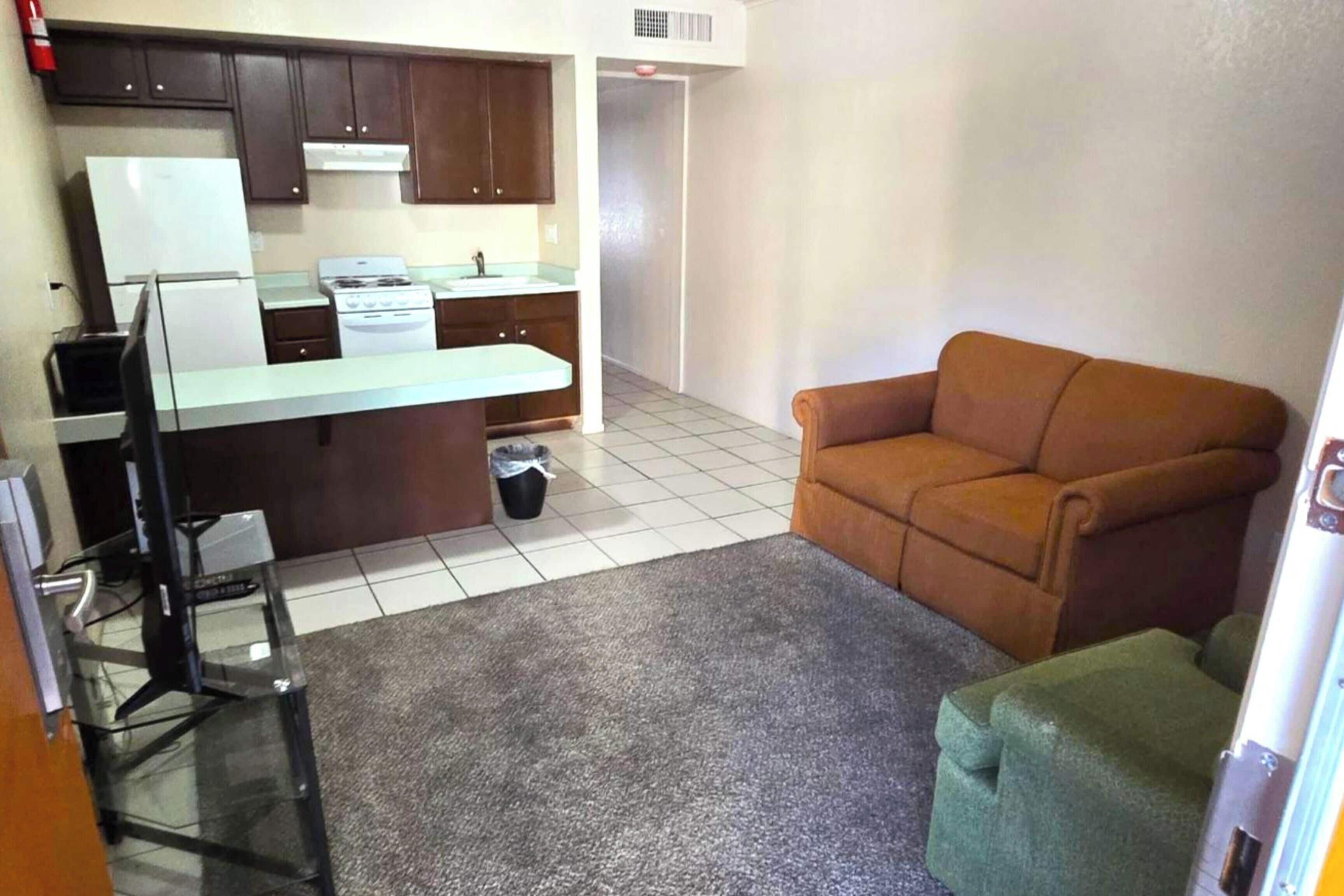 A compact kitchen with a refrigerator, stove, and sink, a brown sofa, and a small green stool.