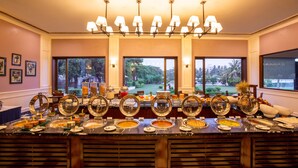 A buffet with multiple food stations, a chandelier, and large windows with a view of greenery. - Sun N Sand Shirdi (Rahata)