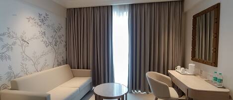 Una habitación de hotel moderna con sofá, una mesita redonda, un escritorio con silla y un ventanal con cortinas.