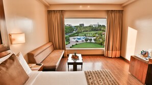 A hotel room with a bed, a sofa, a small table with fruit, a desk, and a view of a park and buildings. - Sun N Sand Shirdi (Rahata)