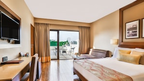 A hotel room with a large bed, a desk, a chair, a TV, and a balcony with a table and chairs. - Sun N Sand Shirdi (Rahata)