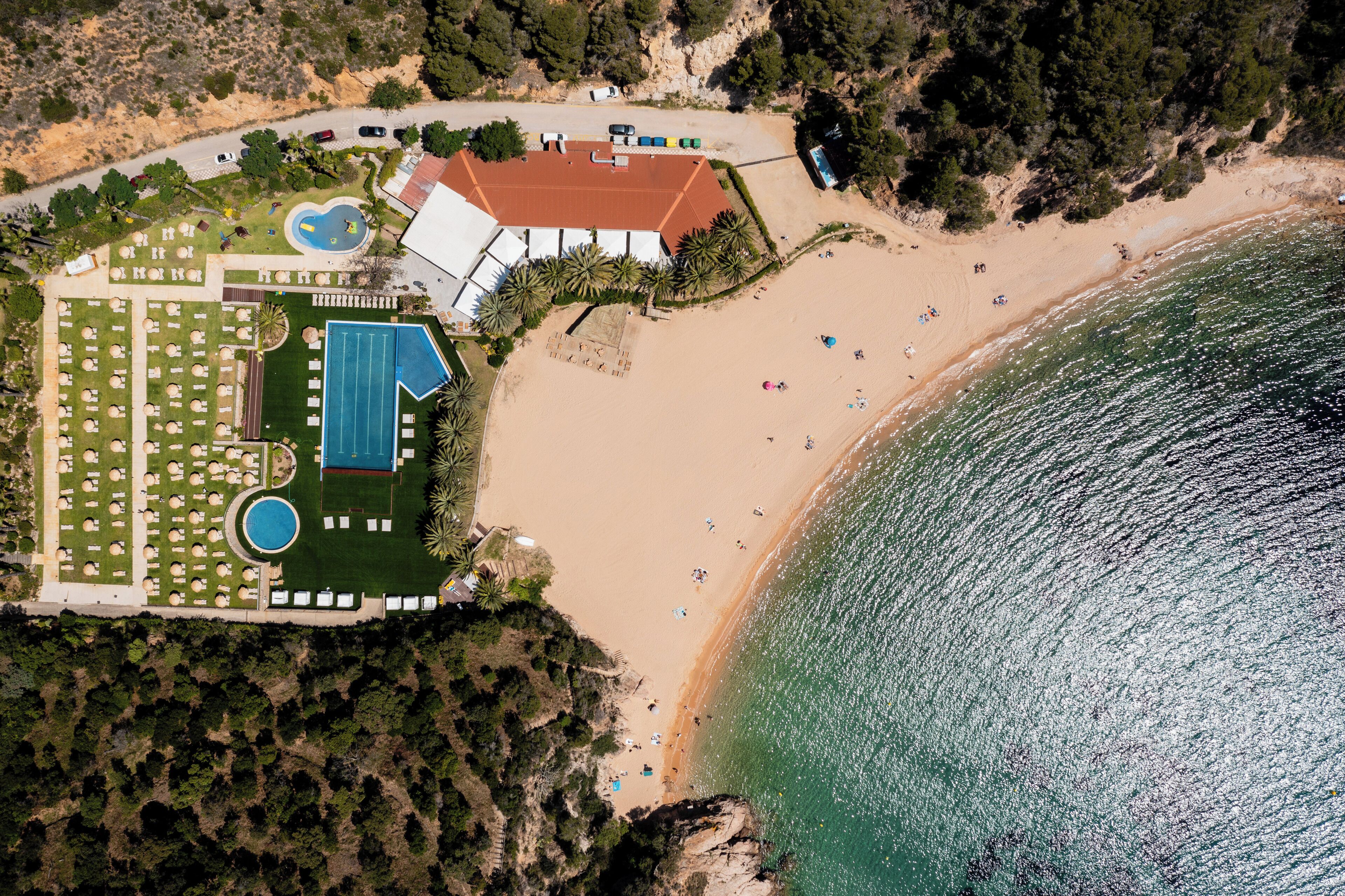 An aerial view of a coastal resort with a sandy beach, a swimming pool, and a tennis court.