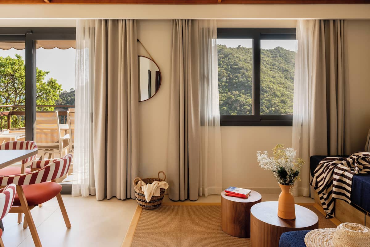 Ein modernes Hotelzimmer mit einem großen Fenster, das Blick auf einen bewaldeten Hang bietet. Es gibt einen Essbereich mit gestreiften Stühlen und einem Tisch, ein Sofa mit einer gestreiften Tagesdecke sowie einen kleinen runden Tisch mit einer Blumenvase.