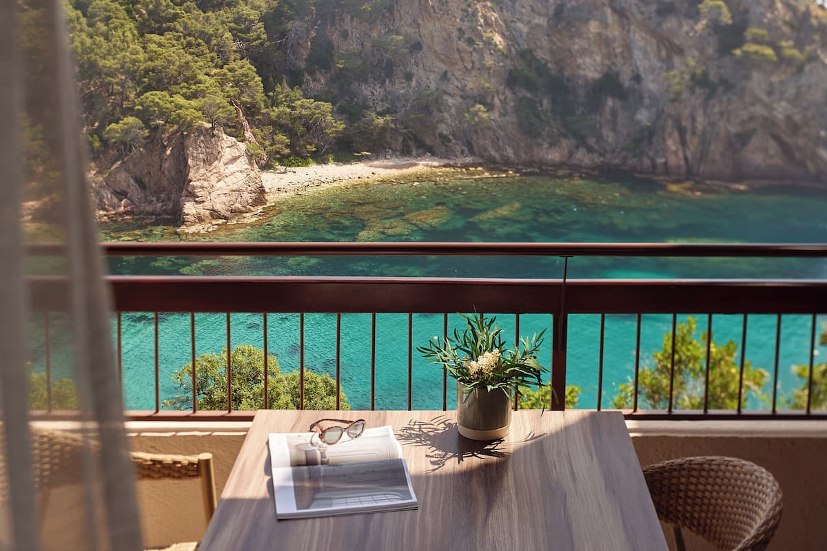 Ein Balkon mit Blick auf eine Küstenlandschaft, ein Tisch mit Pflanze und Brille sowie ein Korbstuhl.