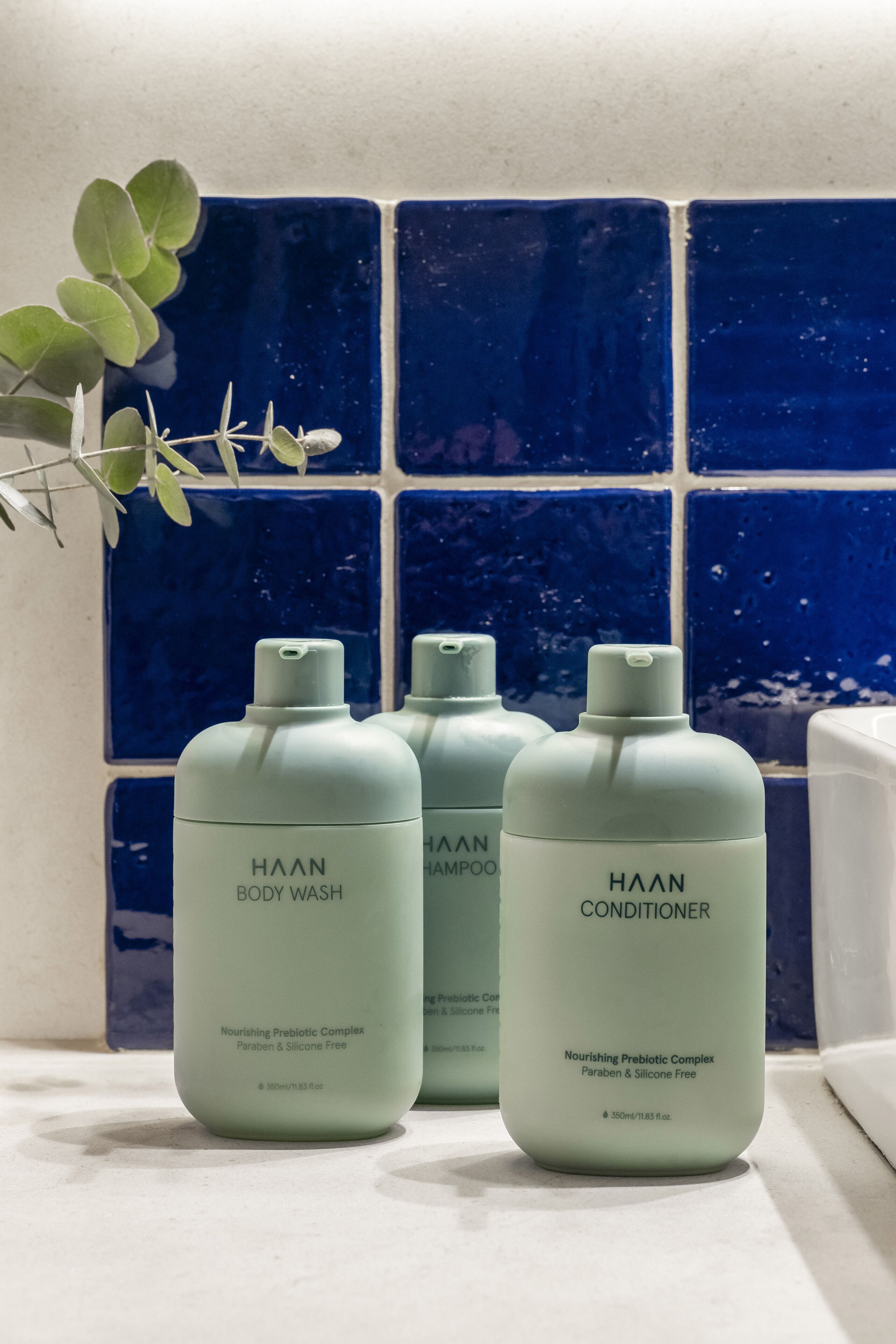 Tre flaconi di bagnoschiuma, shampoo e balsamo Haan su una superficie bianca, con un muro piastrellato blu sullo sfondo.