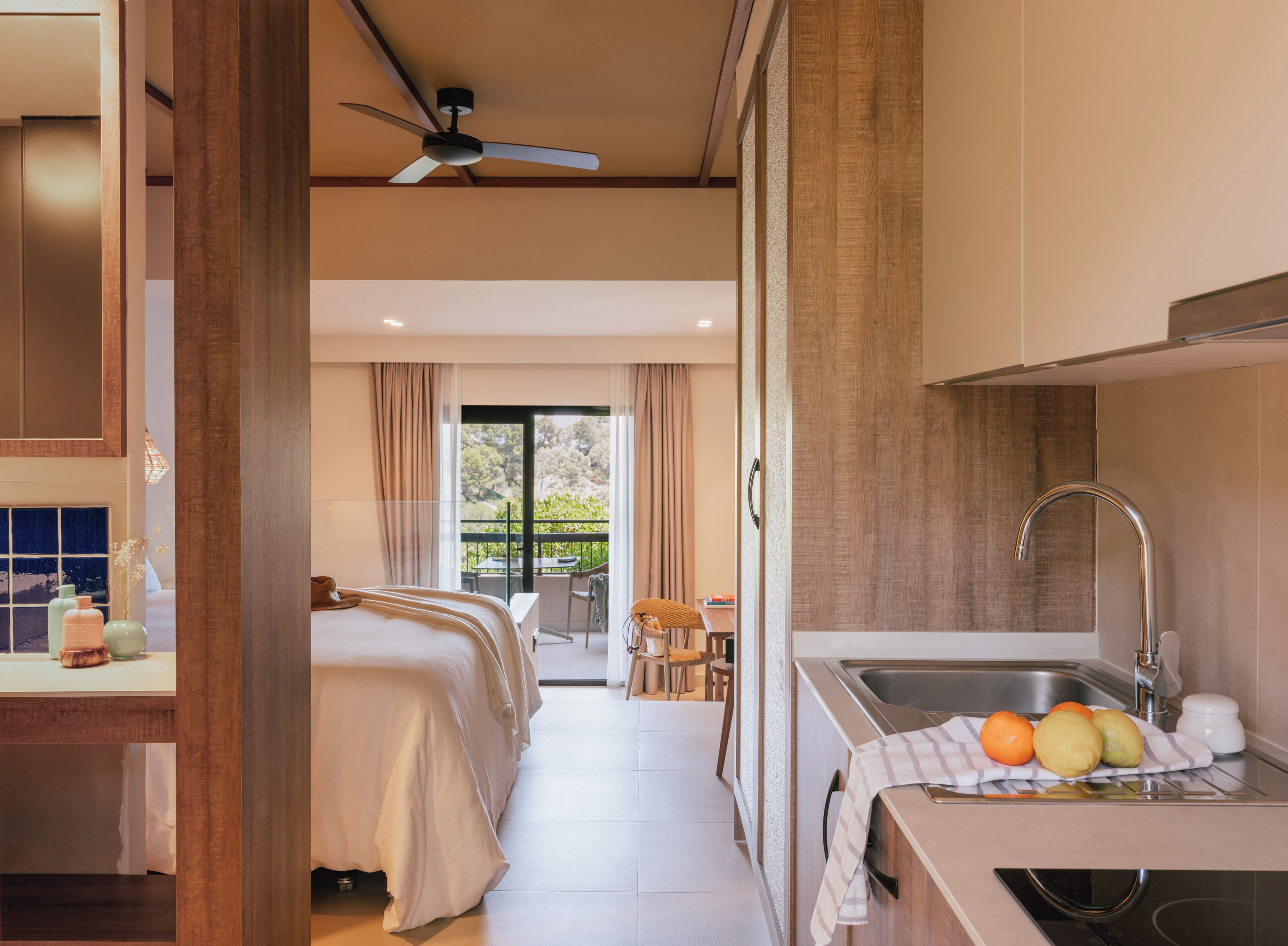 Quarto de hotel com cama, ventilador de teto, varanda com mesa e cadeiras, e área de cozinha com pia e frutas.