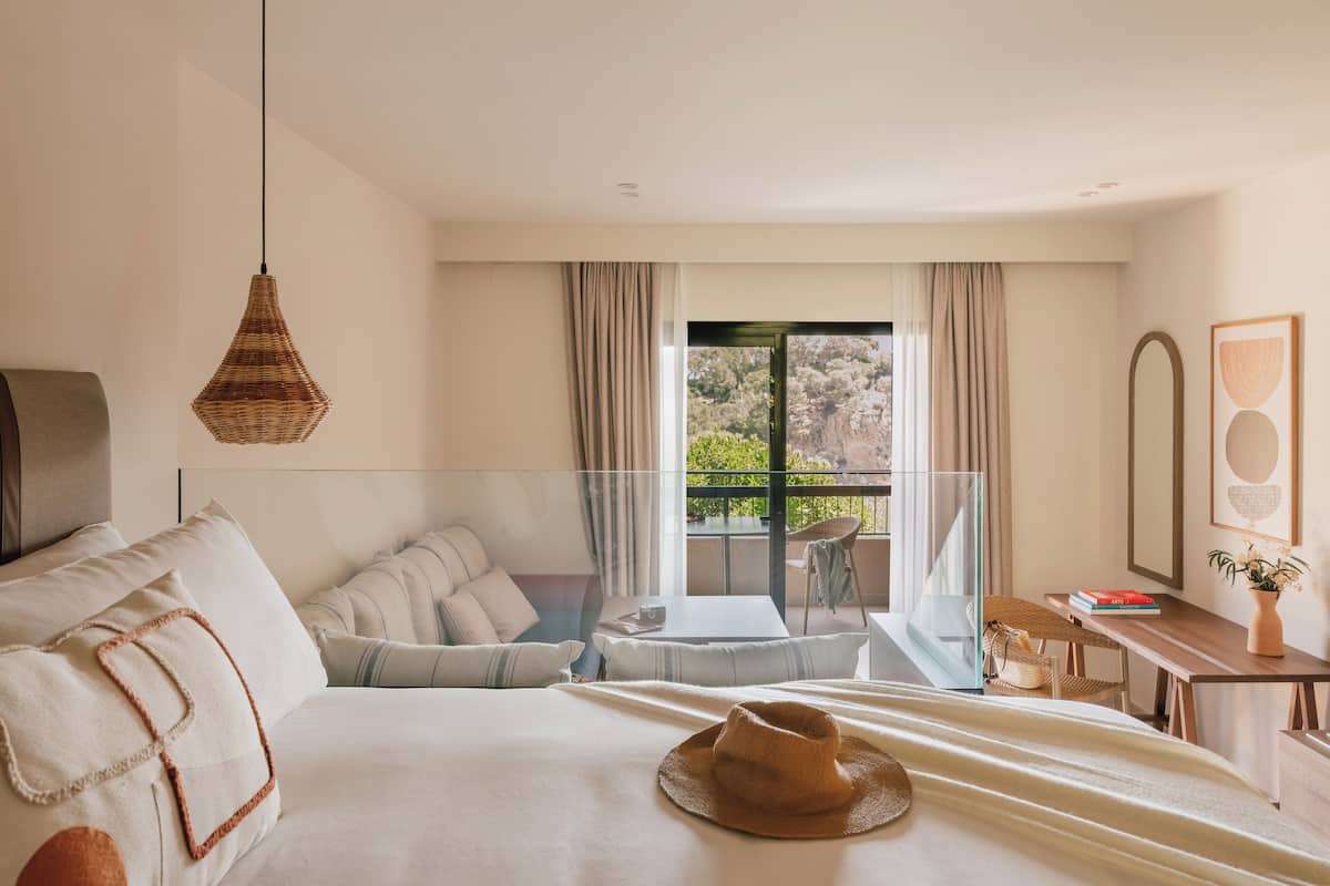 Ein Hotelzimmer mit Bett, einer Sitzecke mit Tisch und Stühlen, einem Balkon mit Blick auf Bäume und einer Pendelleuchte.