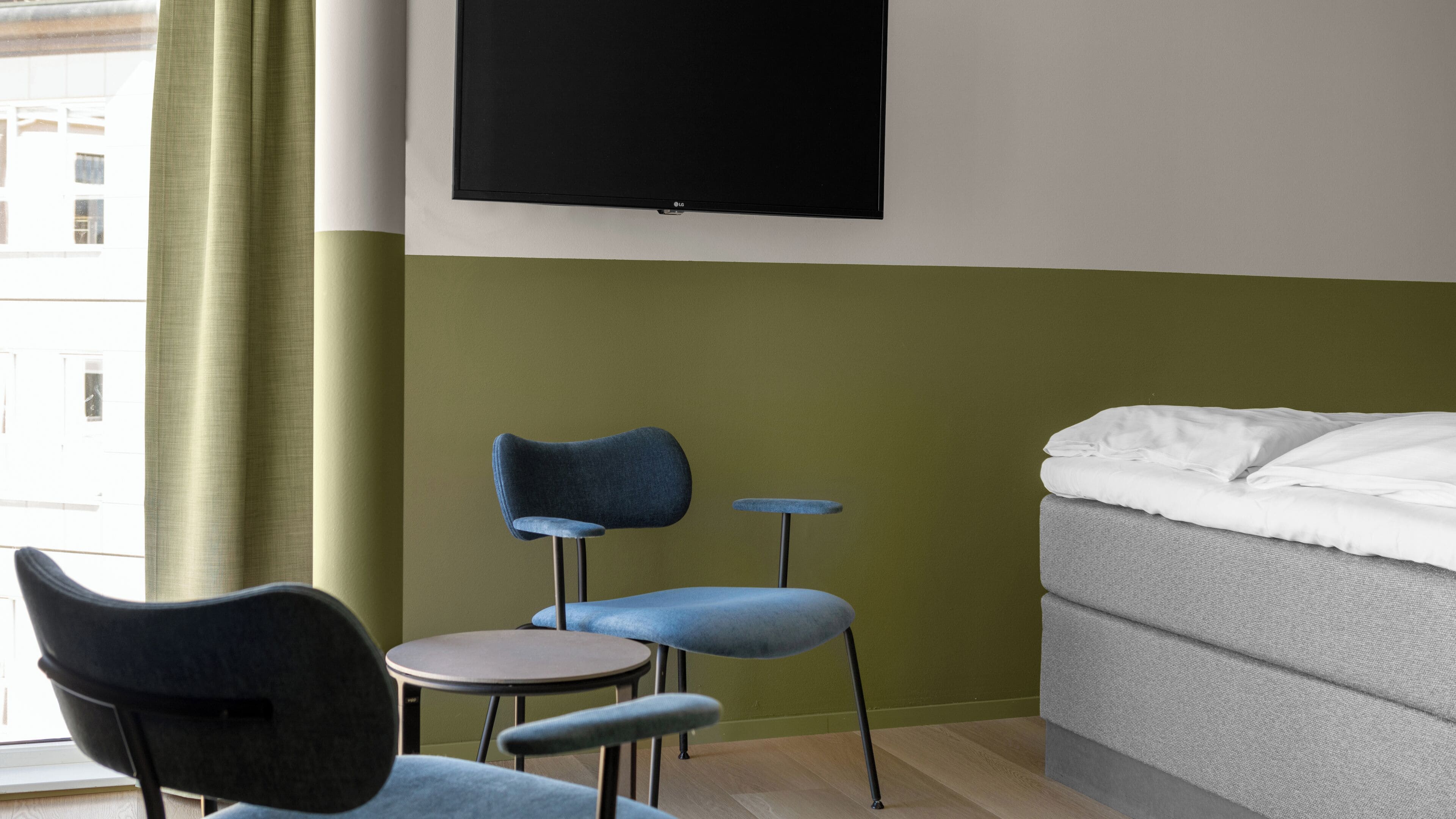 Una habitación de hotel con una cama, dos sillas, una mesita y un televisor montado en la pared.