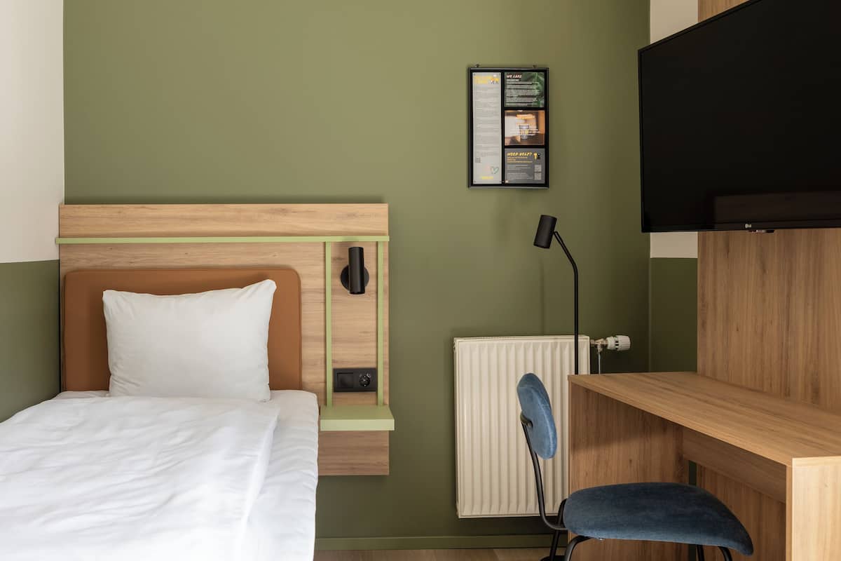 Habitación de hotel con cama, escritorio, silla, radiador y televisor montado en la pared.