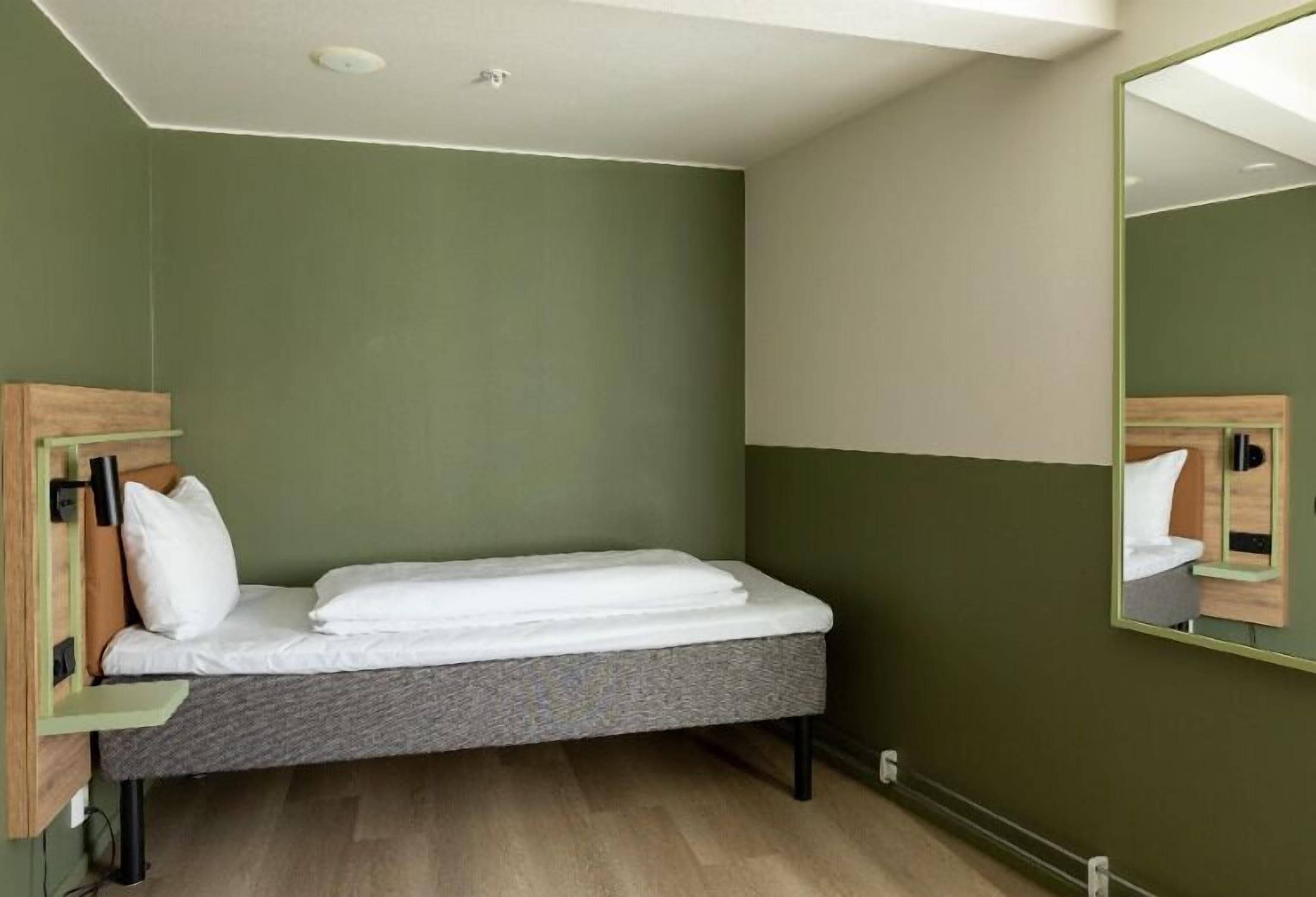 Una habitación pequeña con cama individual, colchón gris, paredes verdes y cabecera de madera.