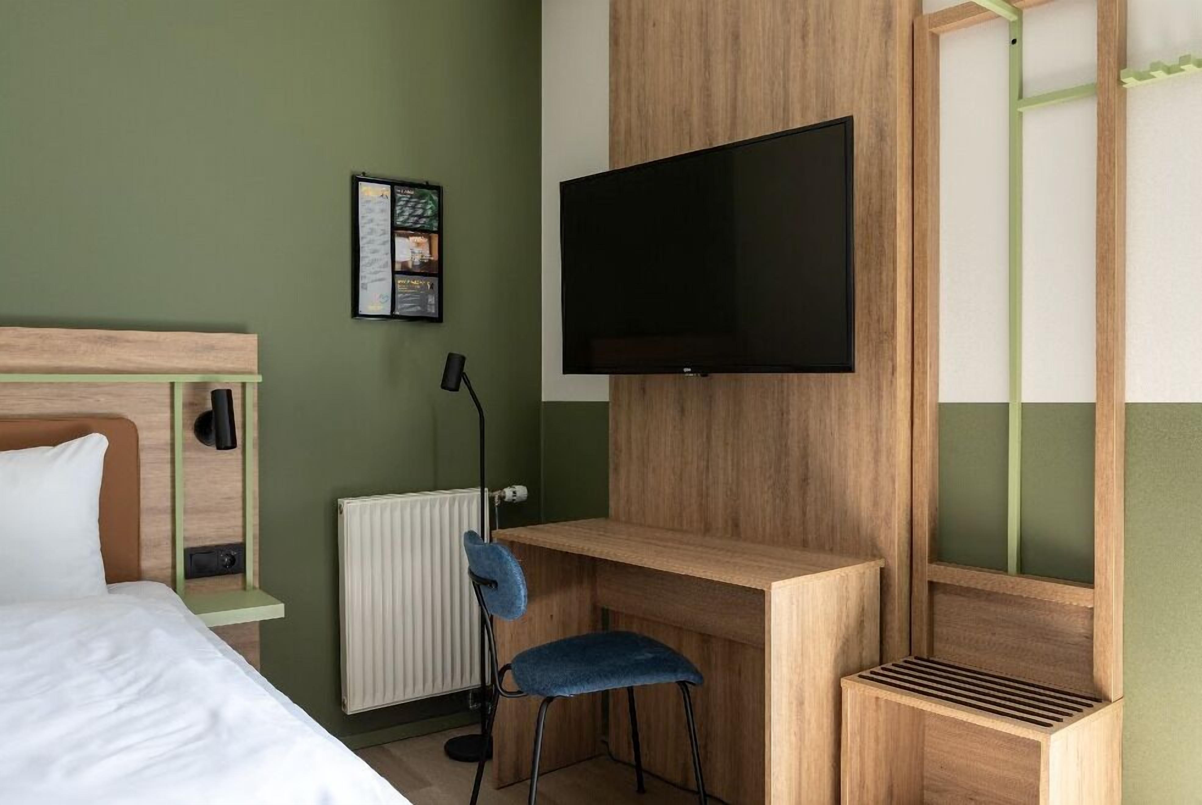 A hotel room with a bed, a desk, a chair, a TV, and a radiator.