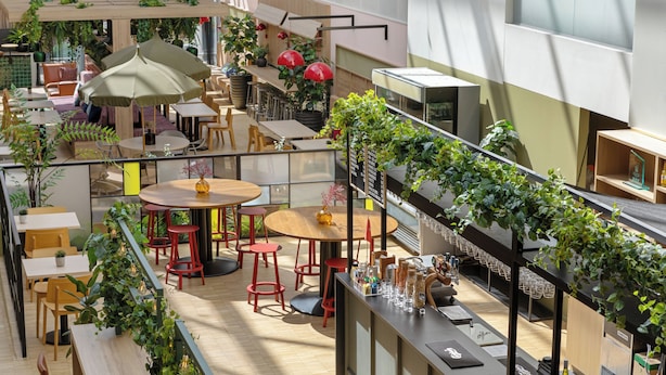 Un espacio interior moderno con varias zonas de estar, incluyendo mesas redondas y taburetes de bar, rodeado de exuberante follaje y plantas en macetas.