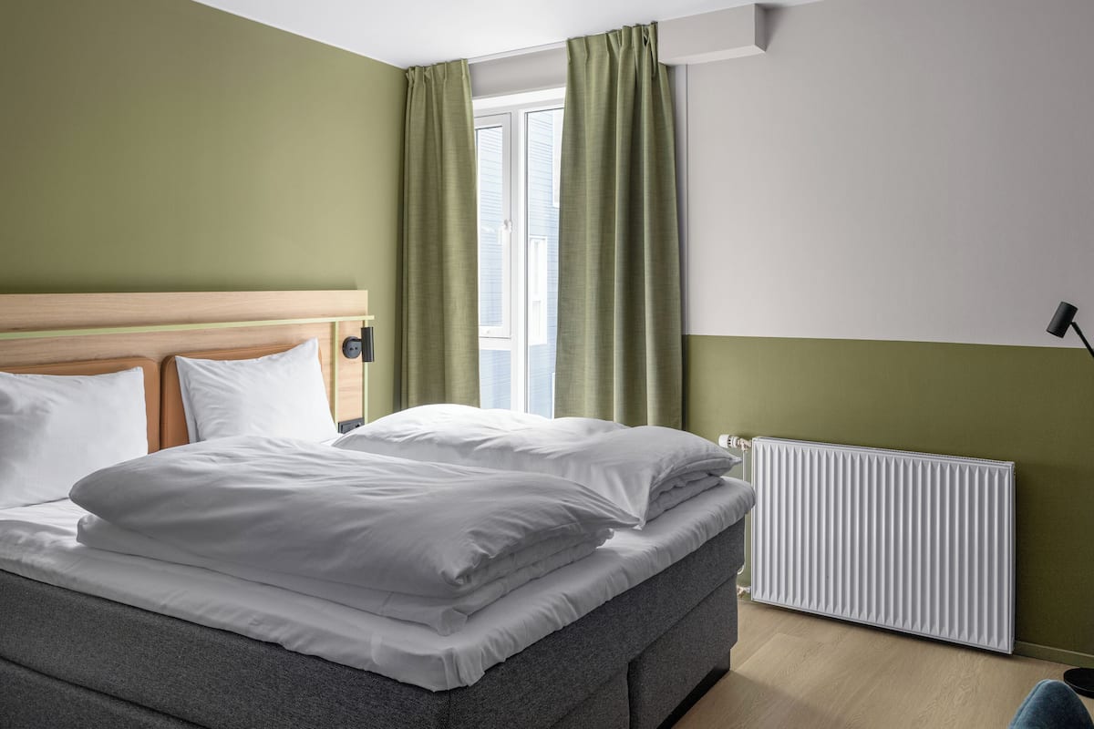 Una cama bien hecha con sábanas blancas, un cabecero tapizado en gris y un radiador en la pared.