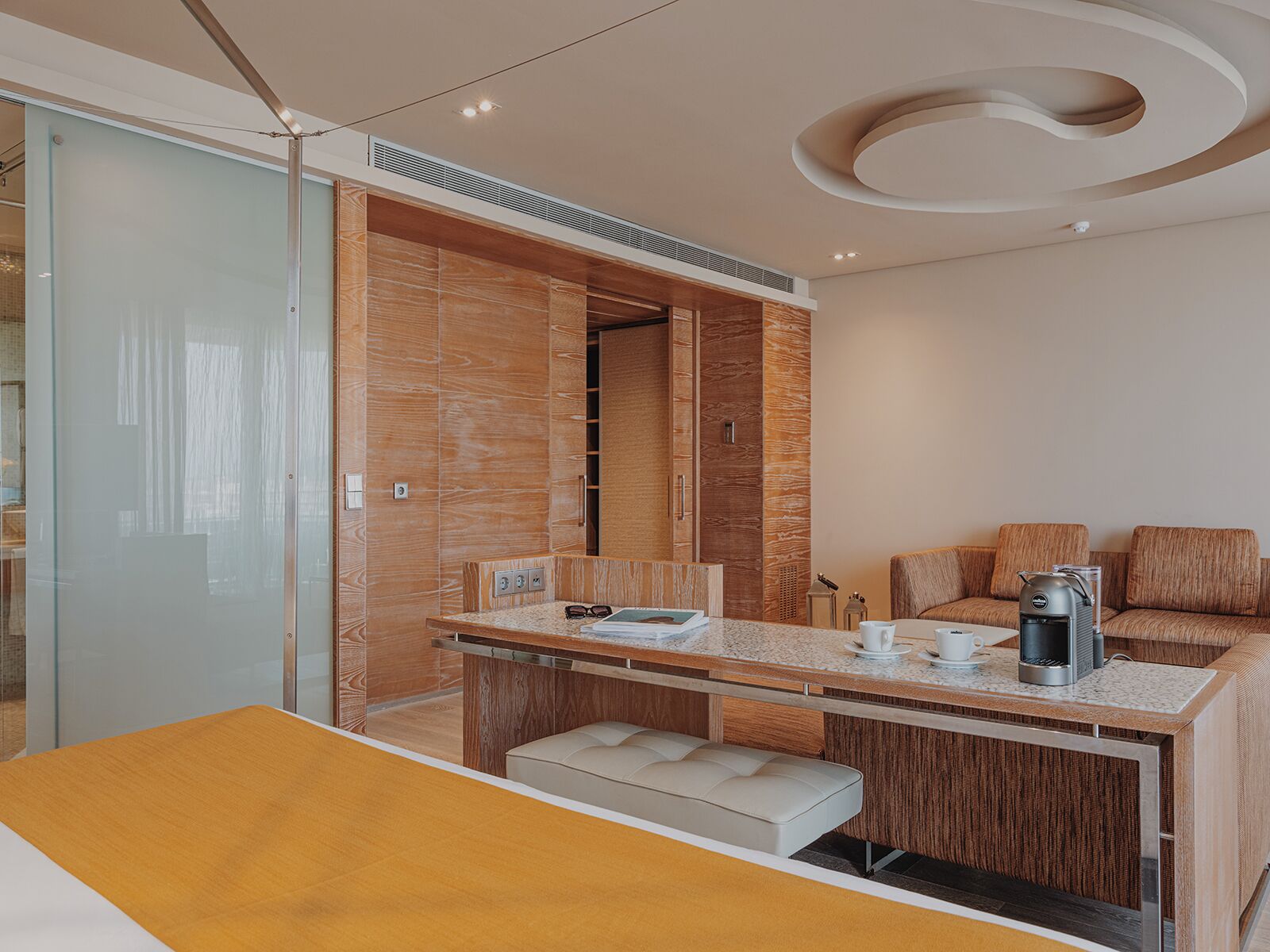 Una habitación de hotel moderna con un tocador de madera, una zona de estar con cojines, una cafetera y una mesita con un bol y vasos.