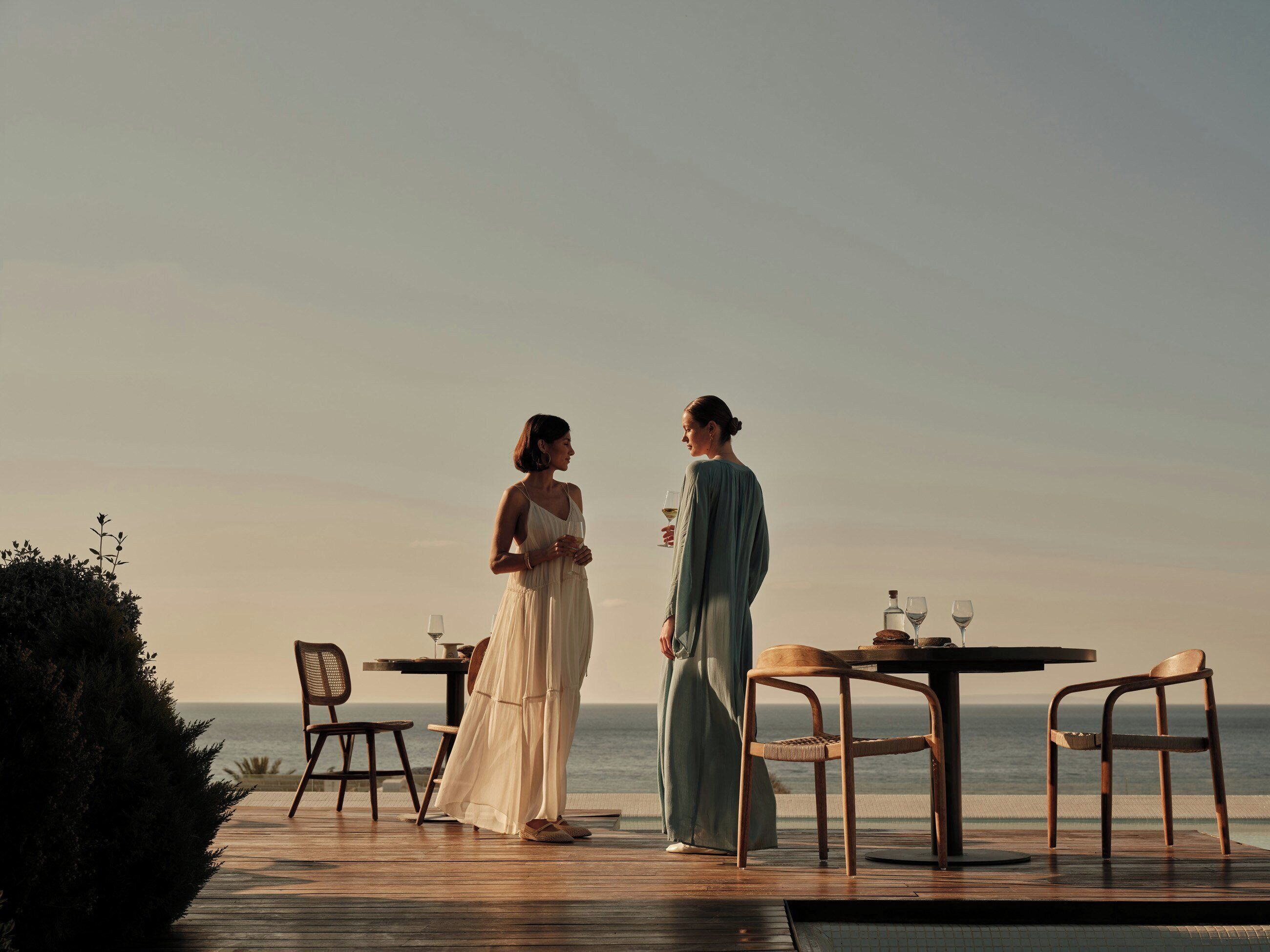 A wooden deck with two people standing, a table with chairs, and a view of the ocean.
