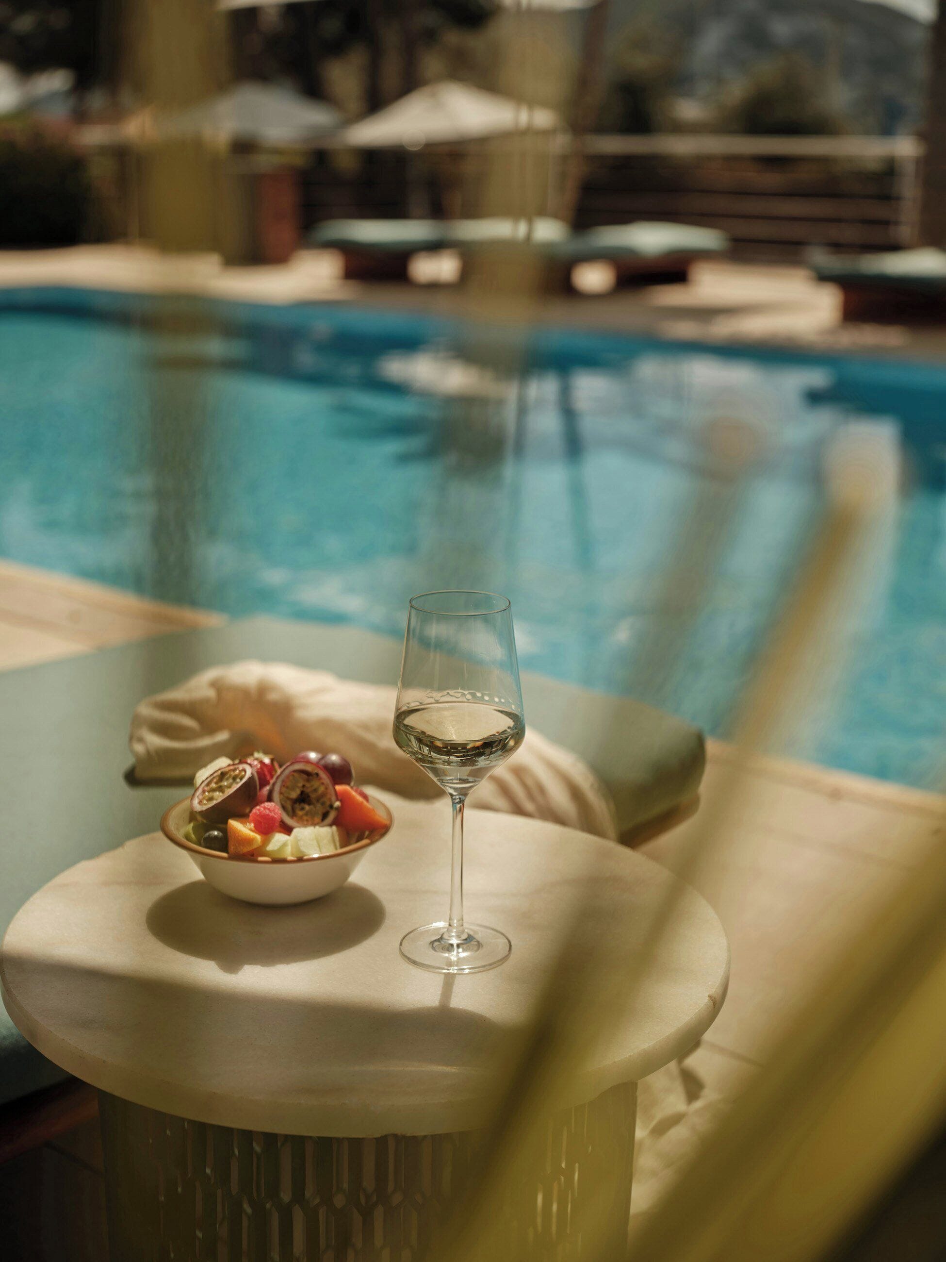 A glass of white wine on a table with a bowl of fruit next to it, overlooking a swimming pool.