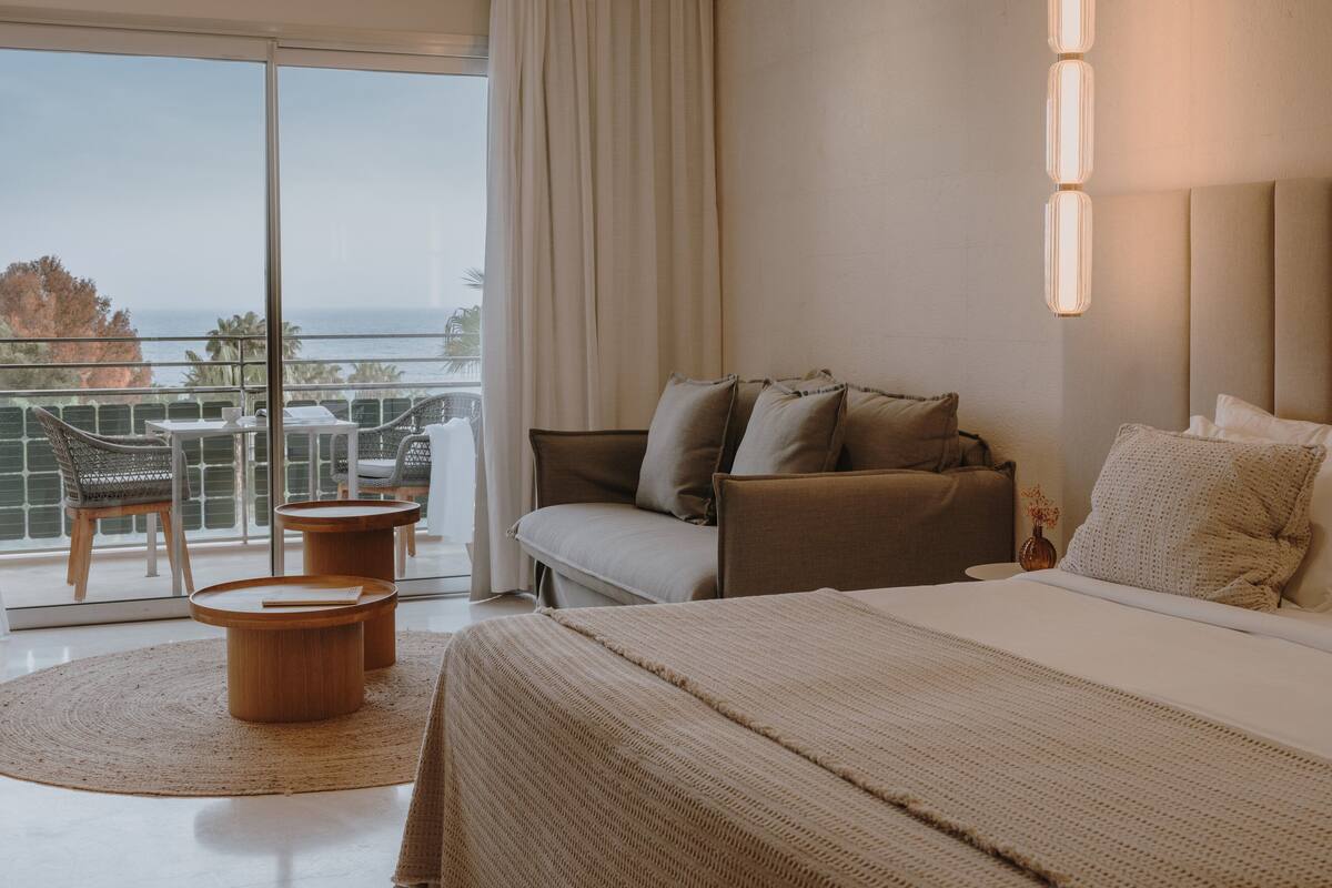Une chambre d’hôtel avec un grand lit, un canapé, un balcon avec une table et des chaises, et une vue sur la mer.