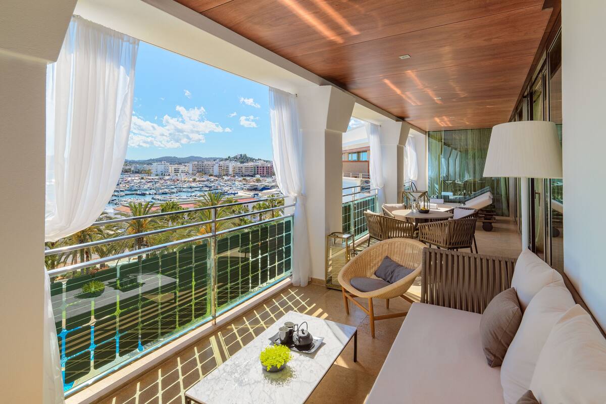 Un balcon offrant une vue sur un port de plaisance et des immeubles, aménagé avec un canapé, des fauteuils et une table sur laquelle est posé un vase de fleurs.