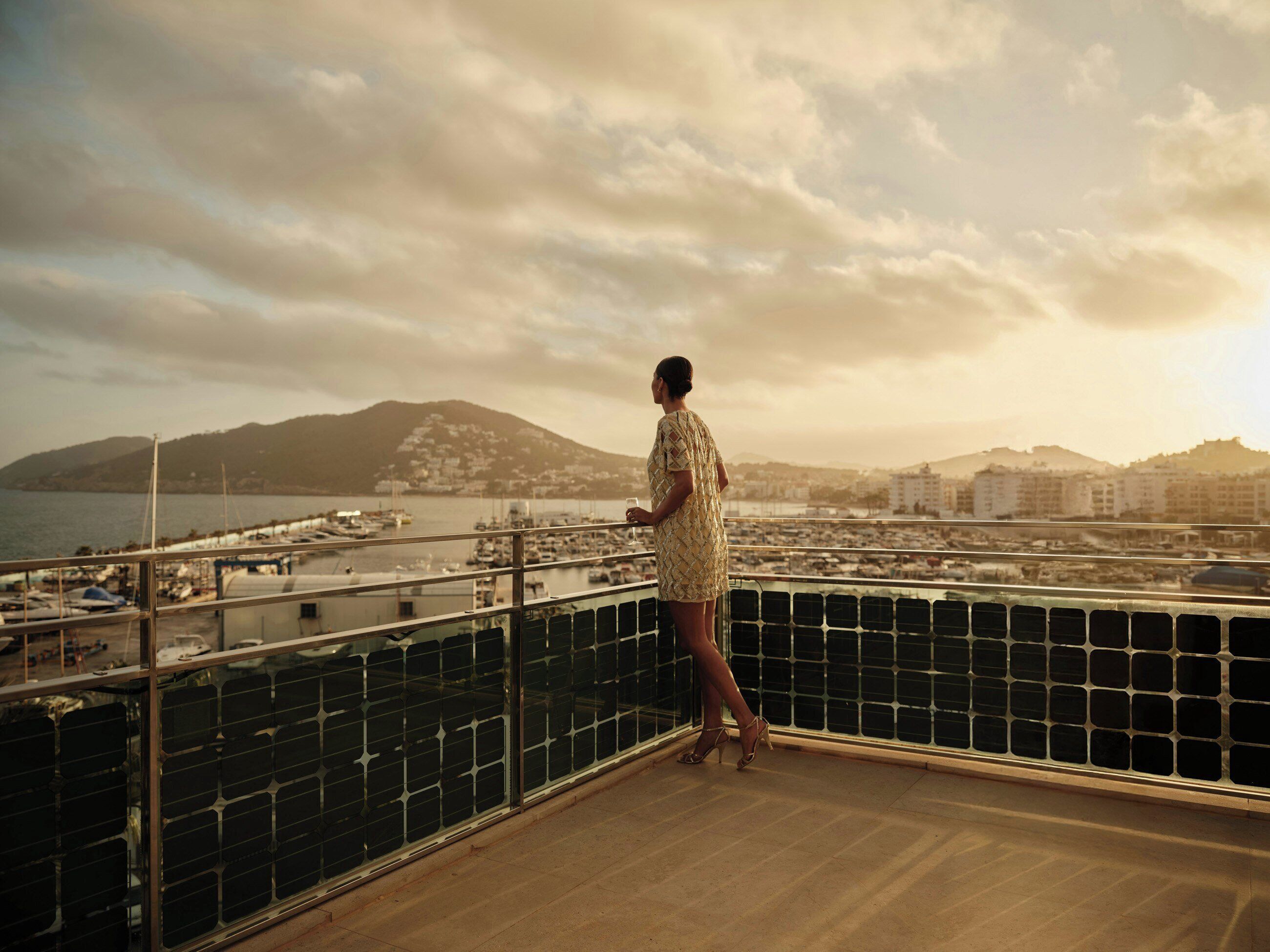 Una persona en un balcón con vista a un puerto deportivo con barcos y edificios al fondo.