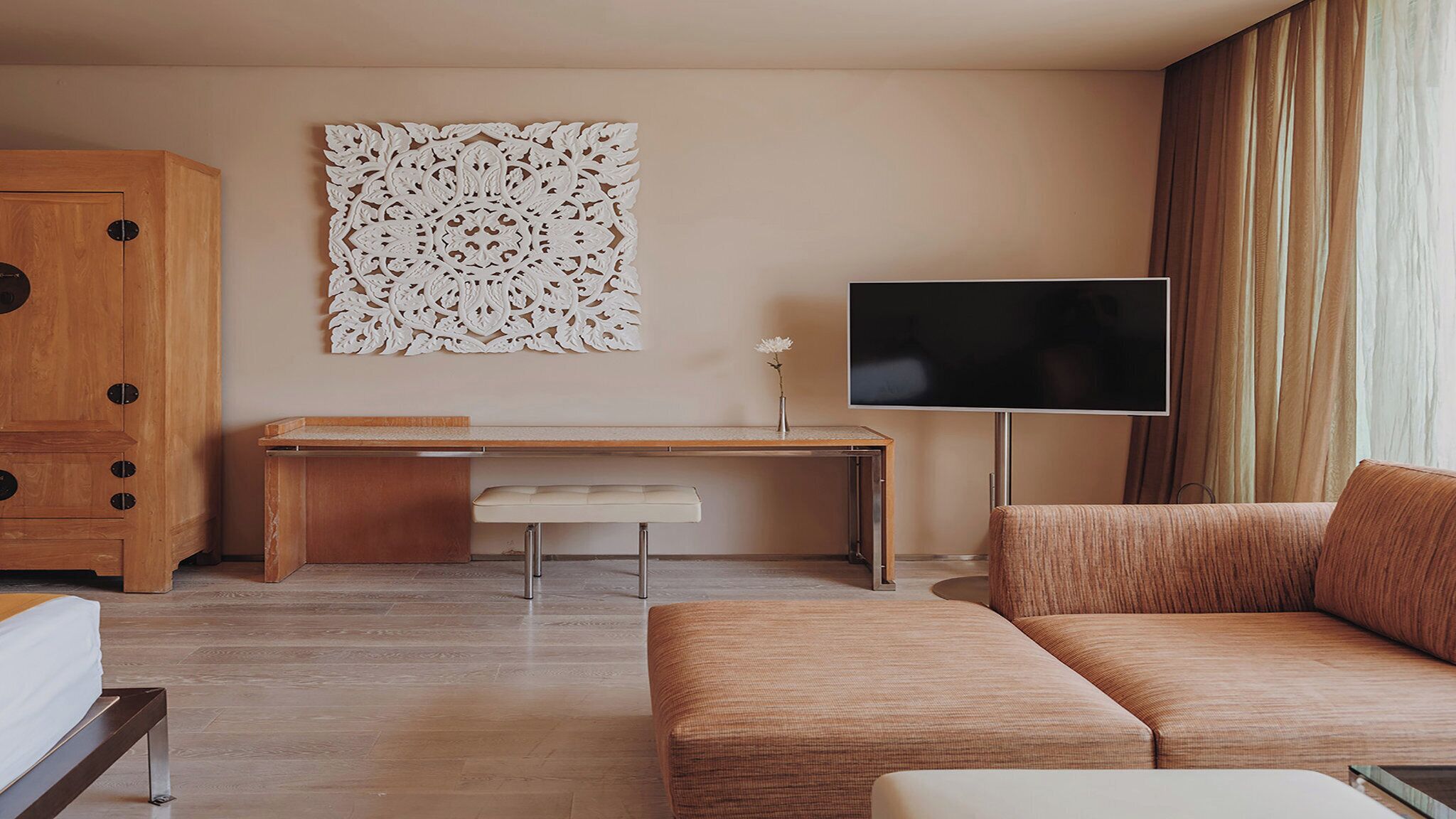 A modern hotel room with a wooden desk, a large flat-screen TV, a cushioned sofa, and a decorative wall piece.