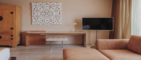 A modern hotel room with a wooden desk, a large flat-screen TV, a cushioned sofa, and a decorative wall piece.
