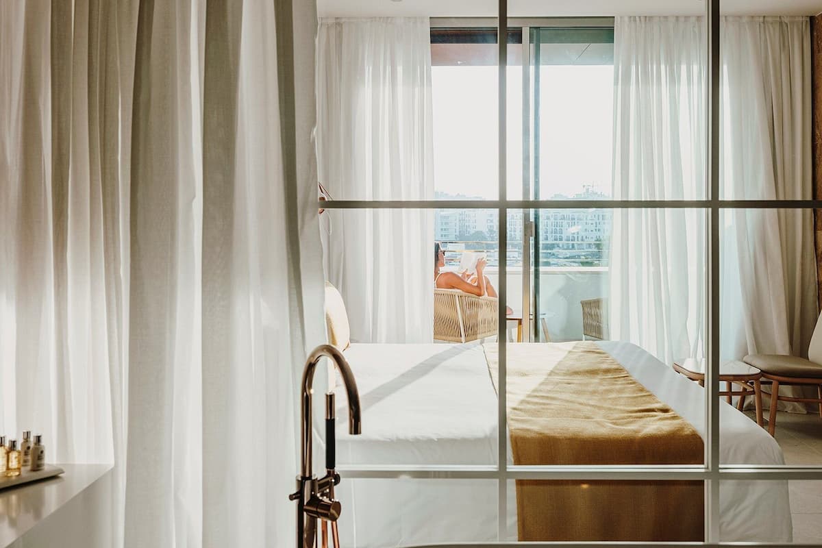 Une chambre d’hôtel dotée d’un grand lit, offrant une vue sur la ville grâce à de larges fenêtres du sol au plafond, et d’un balcon avec des meubles d’extérieur.