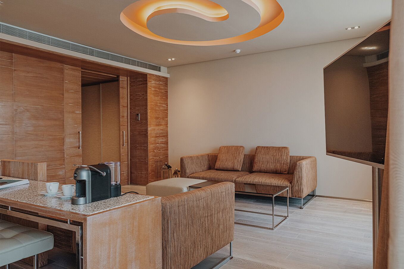 A modern hotel room with a wooden vanity, a sofa, a flat-screen TV, and a coffee machine.