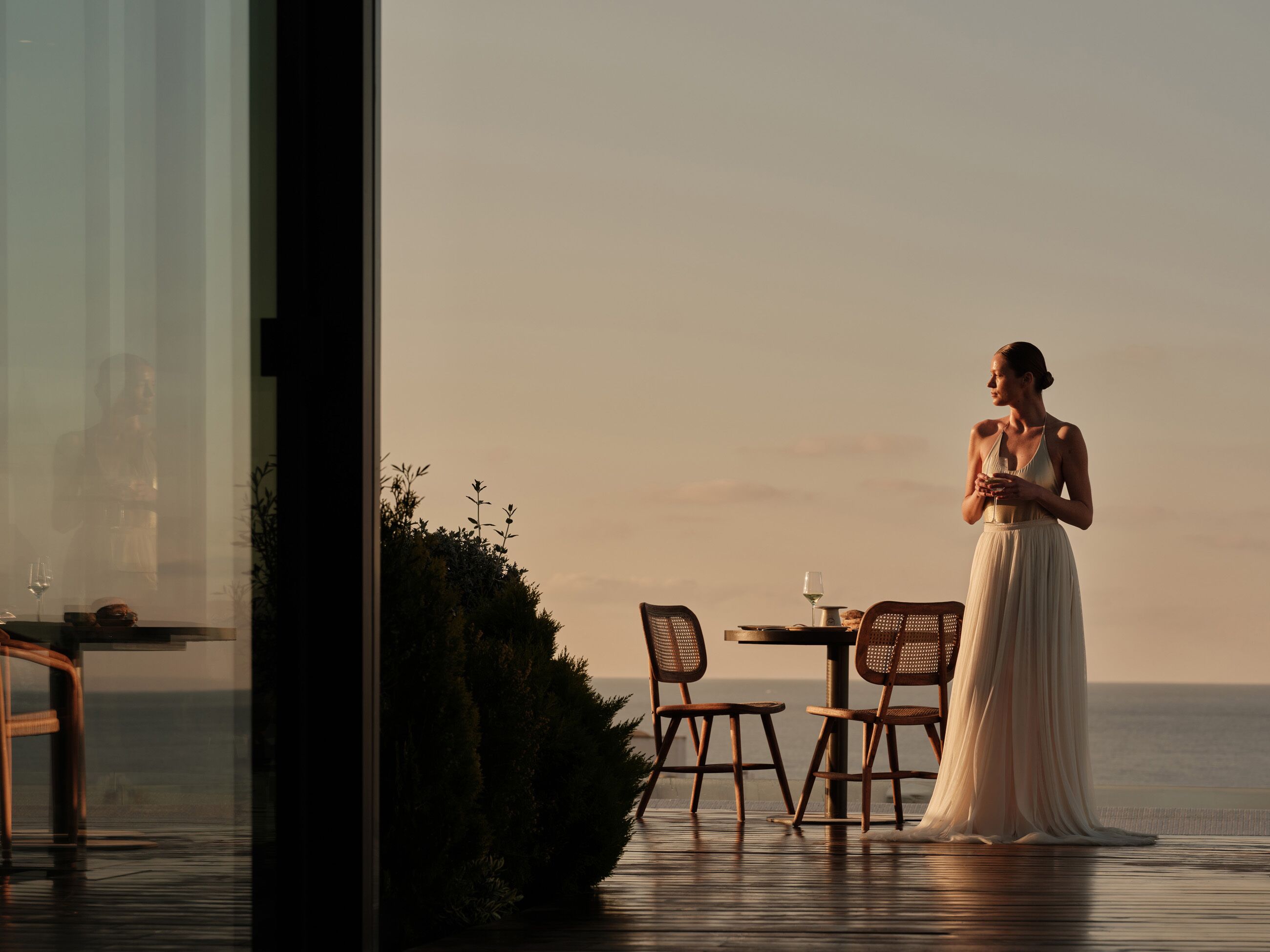 A woman in a white dress standing by a table with chairs, overlooking a body of water.