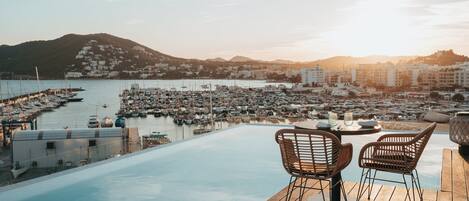 Piscina infinita con terraza de madera y sillas de mimbre, con vistas a un puerto deportivo con barcos y edificios.