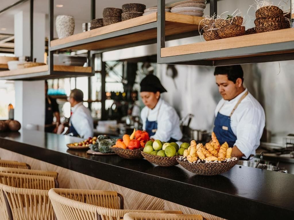 Un área de cocina con chefs preparando comida, repisas con cuencos de frutas y verduras, y sillas de mimbre en primer plano.