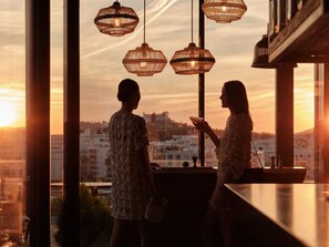 Two people at a bar with a city view, pendant lights, and a clear sky.