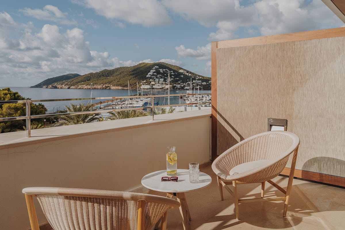 Un balcon agrémenté de fauteuils en osier, d’une petite table et offrant une vue sur une marina et des collines lointaines.
