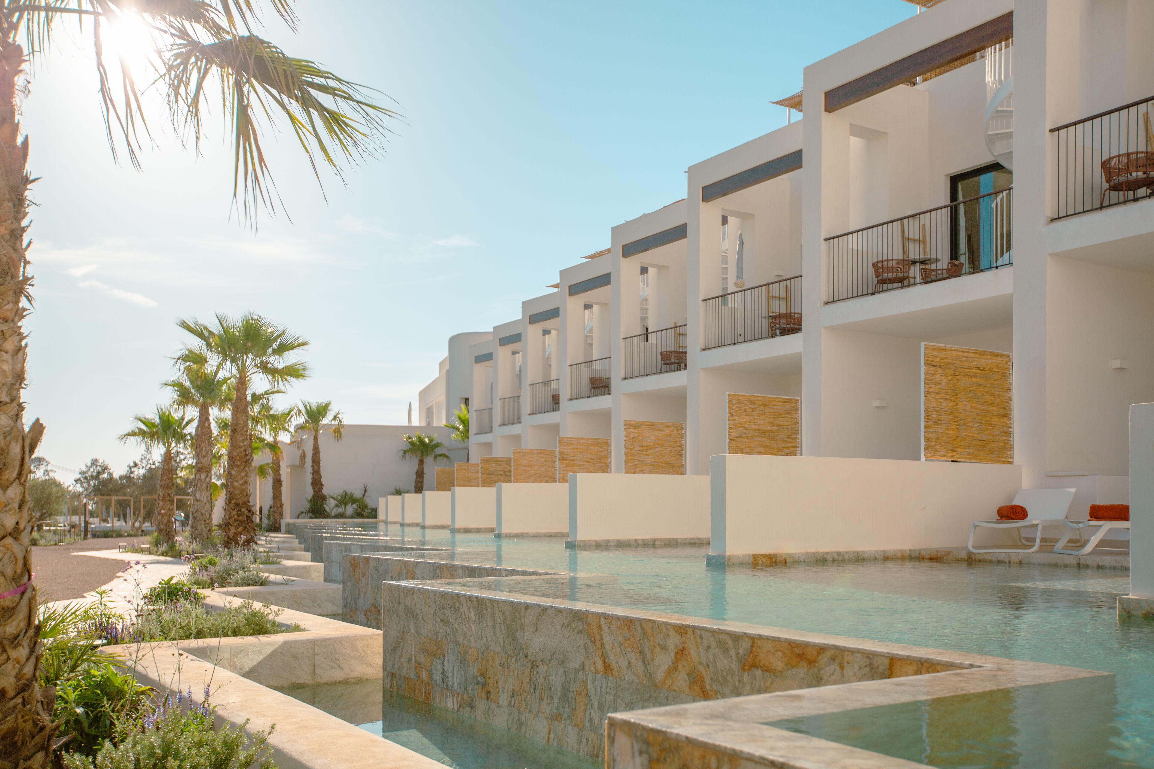 Un hotel moderno con zona de piscina, varios balcones y palmeras.