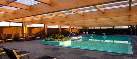 Ein Innenpool mit Holzbalken und Glasverkleidungen, umgeben von Liegestühlen und einer Holzterrasse.