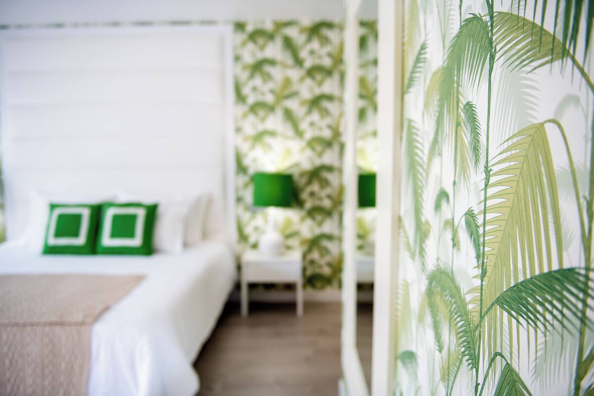 Ein Hotelzimmer mit einem Bett, grünen Kissen, einem Nachttisch, einer Lampe und einer Wand mit Palmenblattmuster.