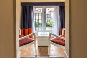 Ein Zimmer mit Blick durch ein Fenster, ausgestattet mit zwei Stühlen mit gemusterten Kissen, einem weißen Hocker und einem Fliesenboden.