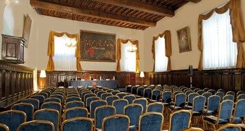 A conference room with rows of blue chairs, wooden paneling, and a large painting on the wall.