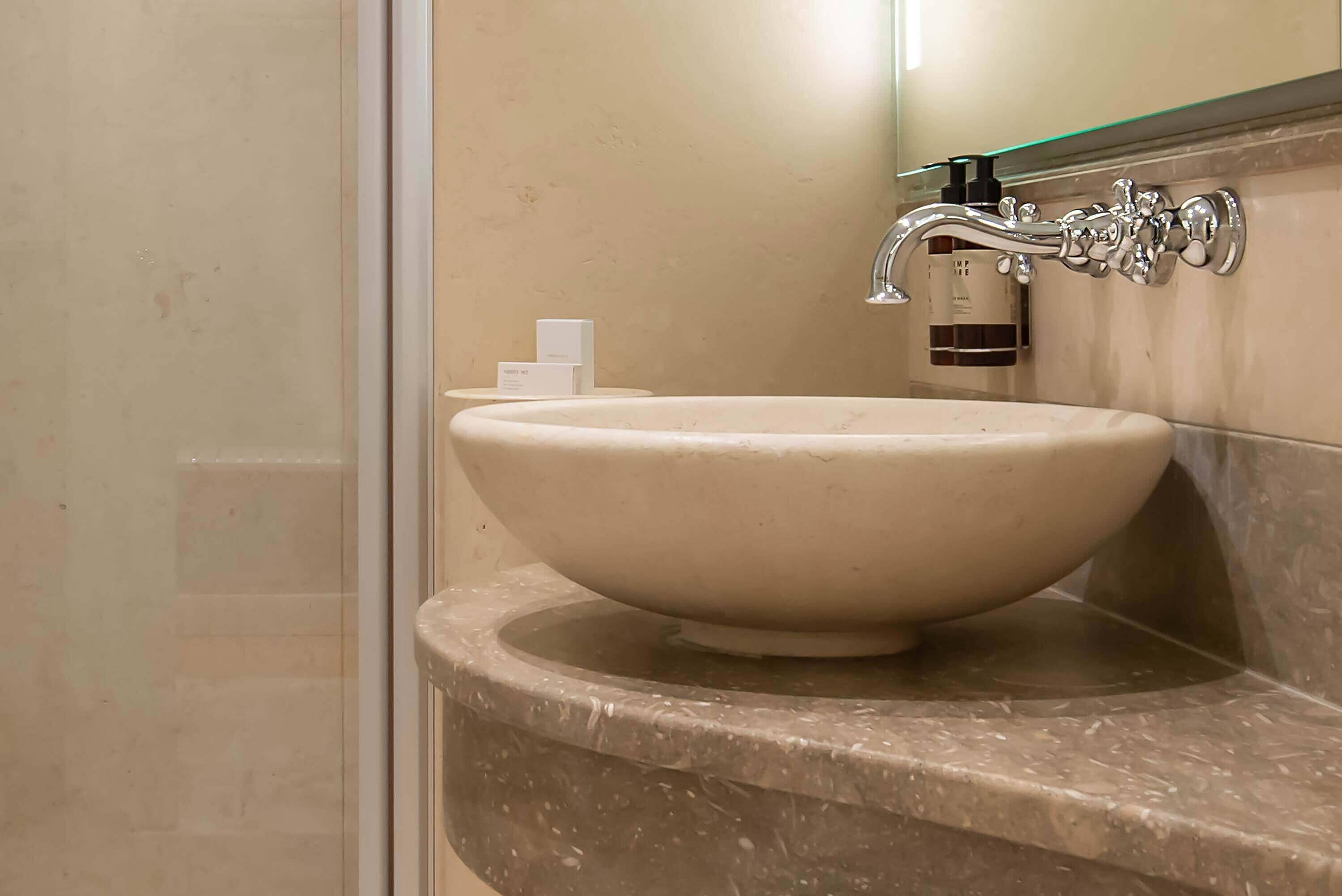 Un baño con un lavabo grande y redondo de color beige sobre una encimera de mármol curvada, un grifo cromado y un box de ducha de vidrio.