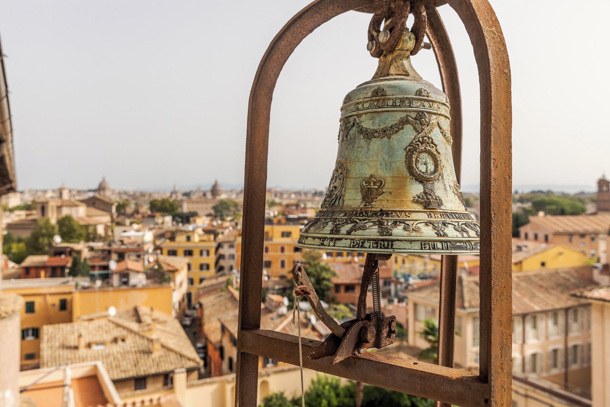 Une cloche ornée d’inscriptions, placée sur un fond représentant un paysage urbain avec des bâtiments et des toits.