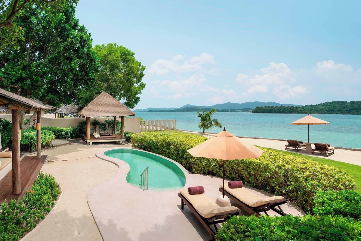 A pool area with lounge chairs and umbrellas, surrounded by greenery and overlooking a body of water.
