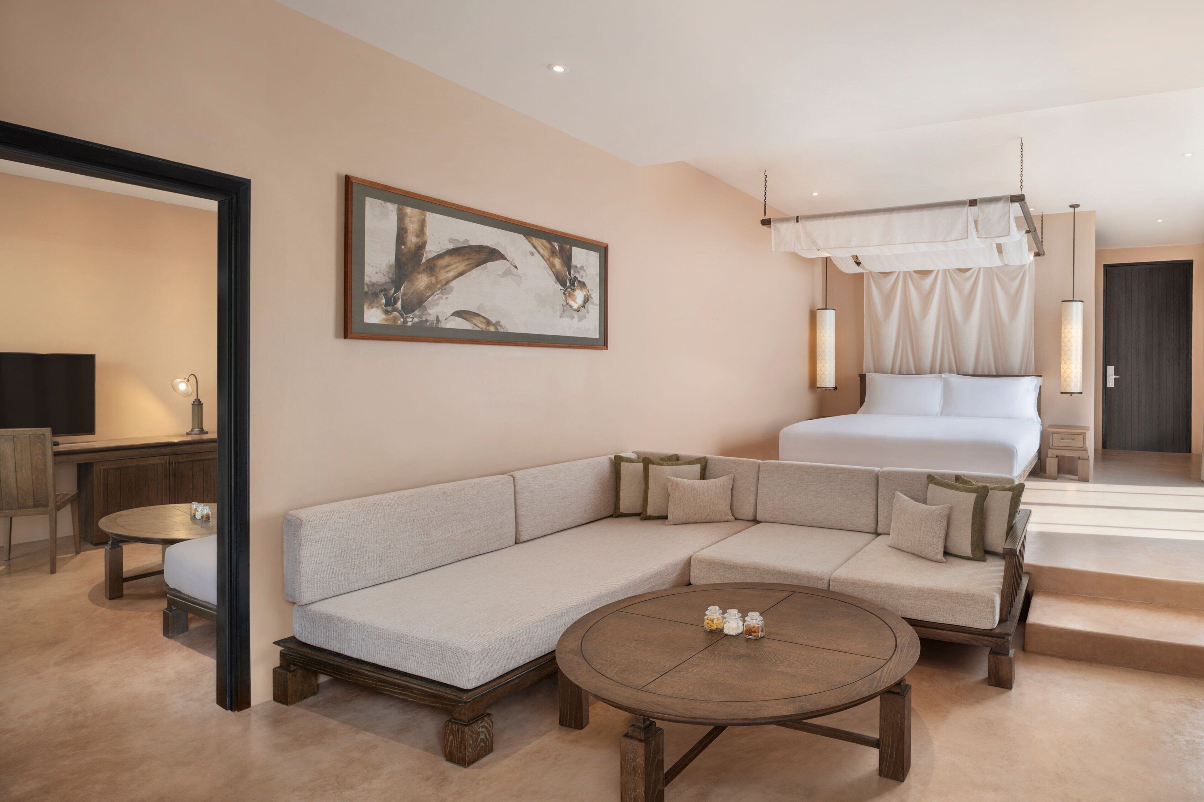 A modern hotel room with a large sofa, a round coffee table, a bed with a canopy, and a painting on the wall.