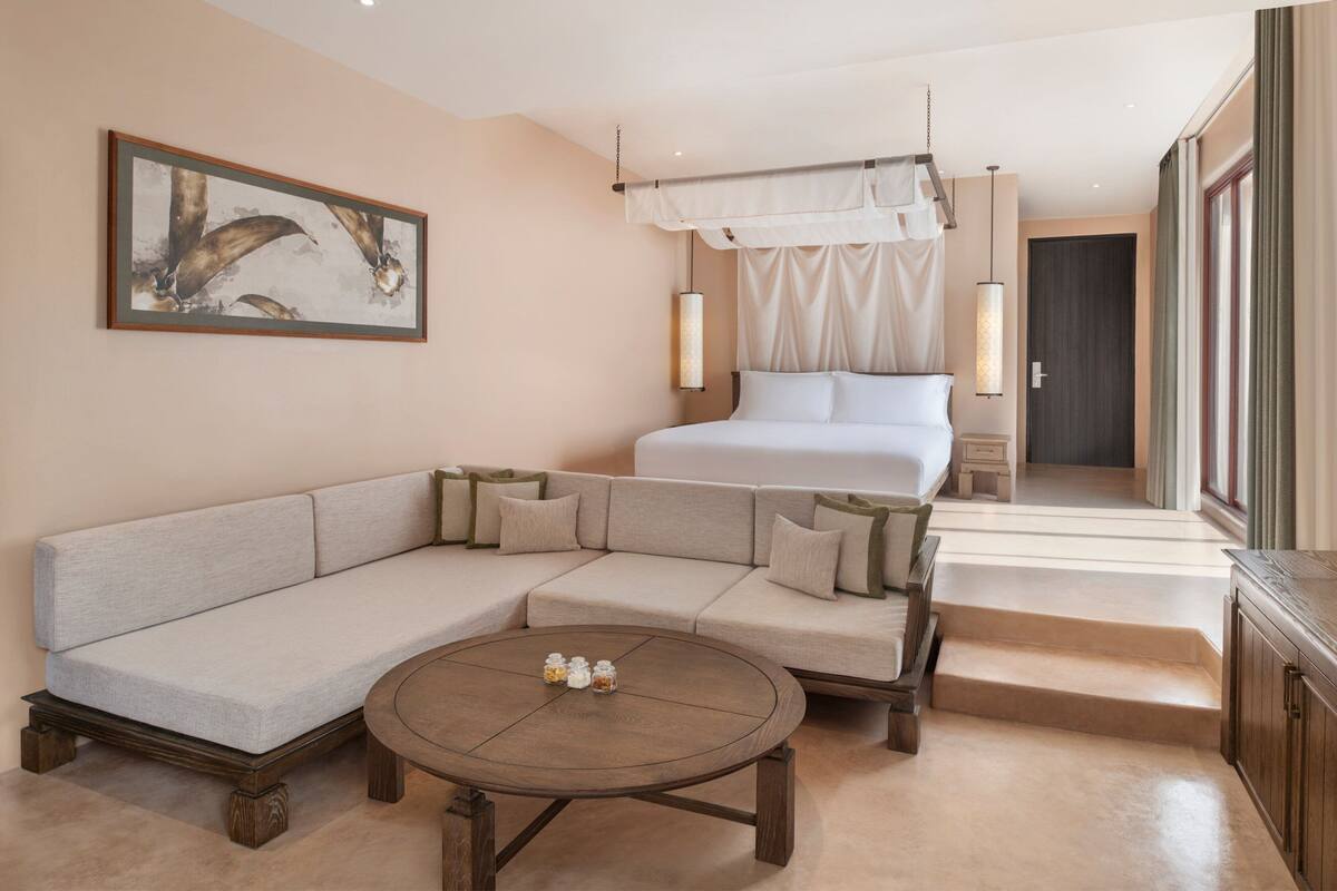A modern hotel room with a sectional sofa, a round coffee table, a bed with a canopy, and a large window with curtains.
