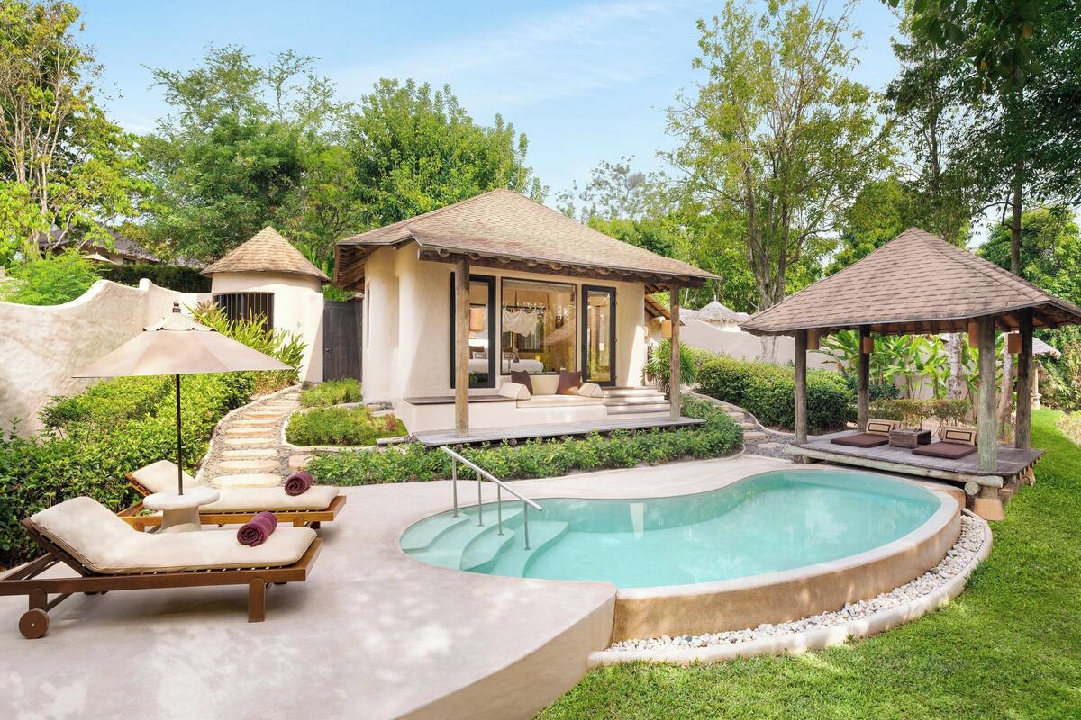 A pool area with lounge chairs, a gazebo, and a small building with a tiled roof.