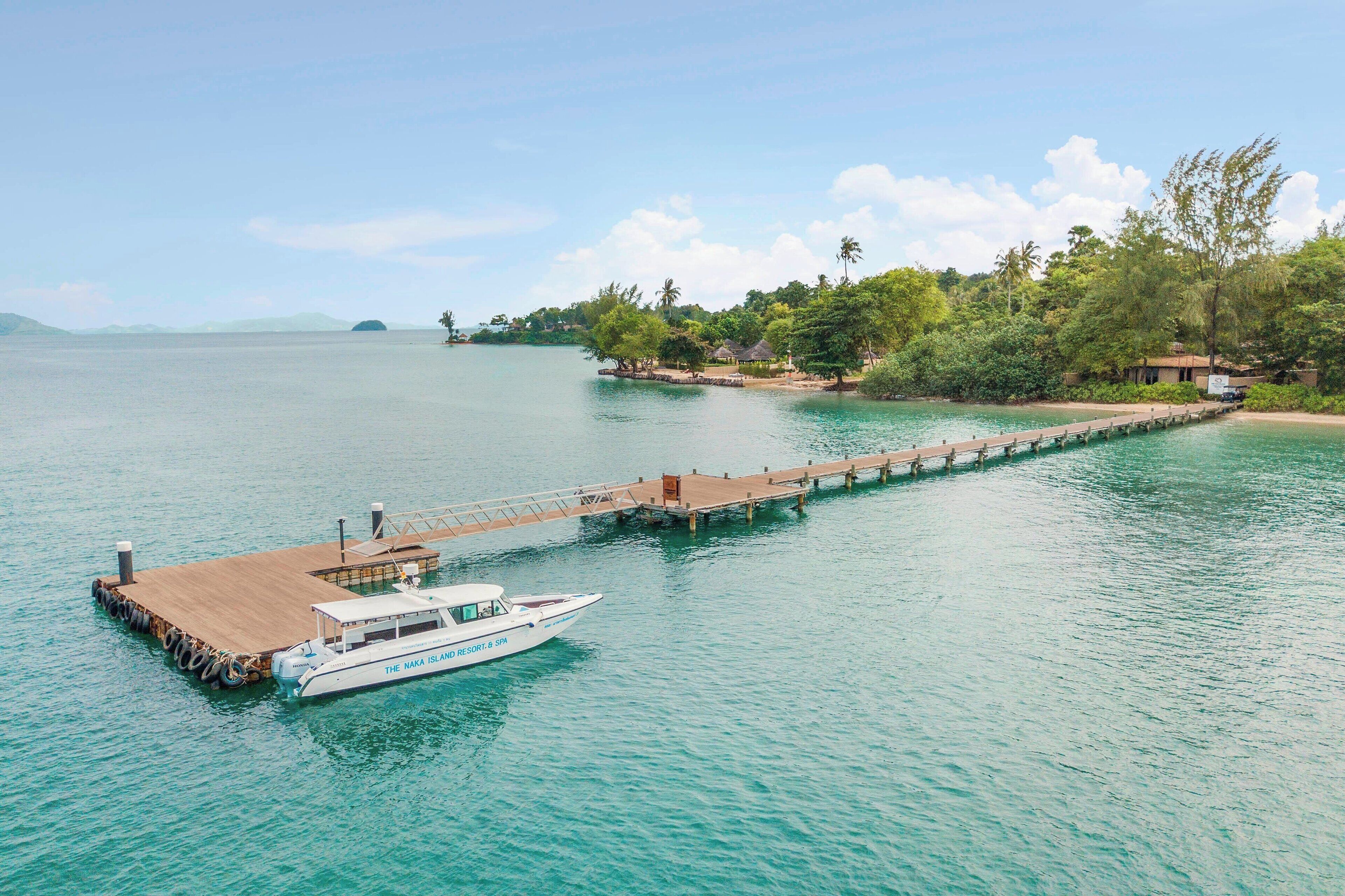 A boat docked at a wooden pier extending into a calm body of water, with a small building and lush greenery in the background.